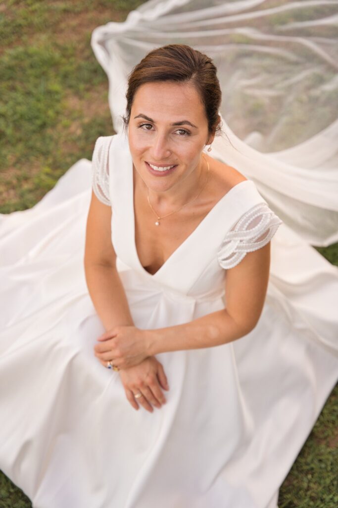 Portrait de la mariée vu d'en haut, souriante, sa robe et son voile étalés dans l'herbe.