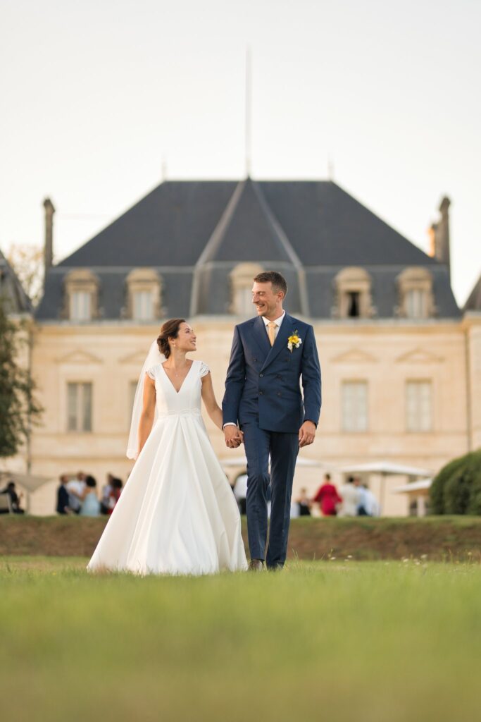 Les mariés marchent main dans la main sur la pelouse, face à la façade du château.