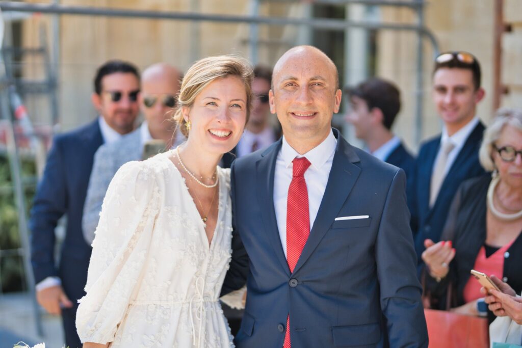 Un homme en costume bleu et cravate rouge et une femme en robe blanche sourient, entourés d'invités.