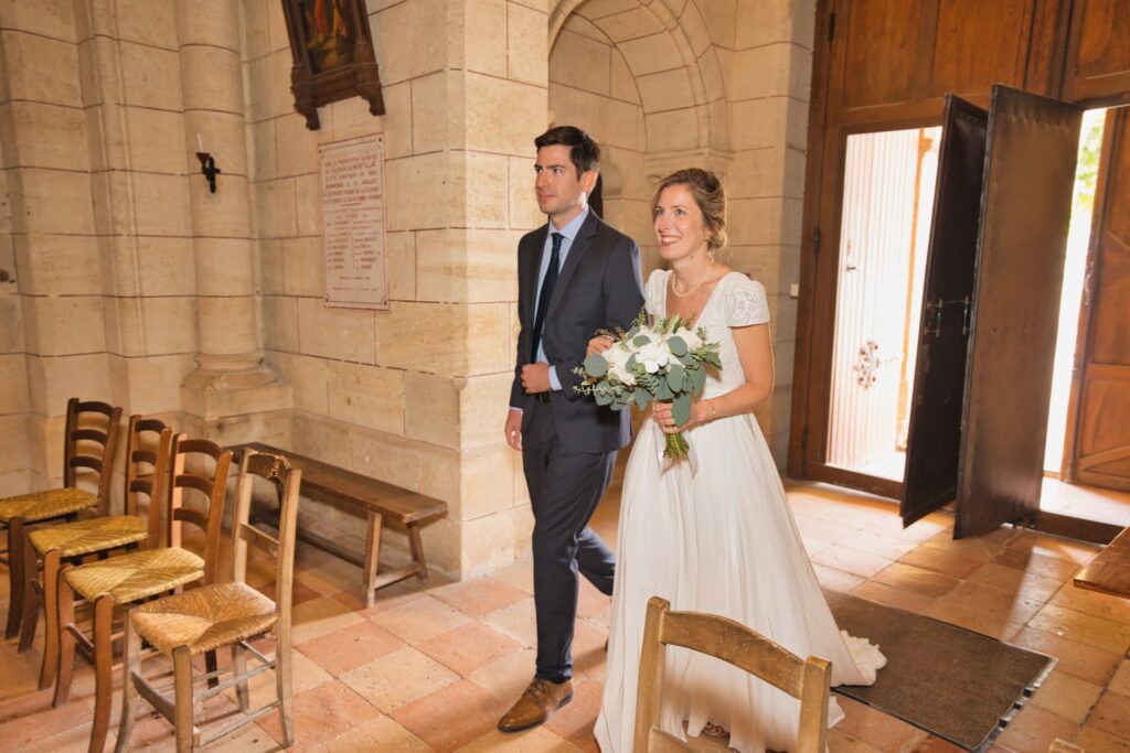 La mariée en robe blanche et un homme en costume sombre entrent dans l'église par une porte ouverte.