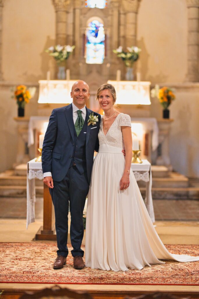 Les mariés sourient face à l'objectif devant l'autel d'une église romane, avec un vitrail et des bouquets de fleurs en arrière-plan.