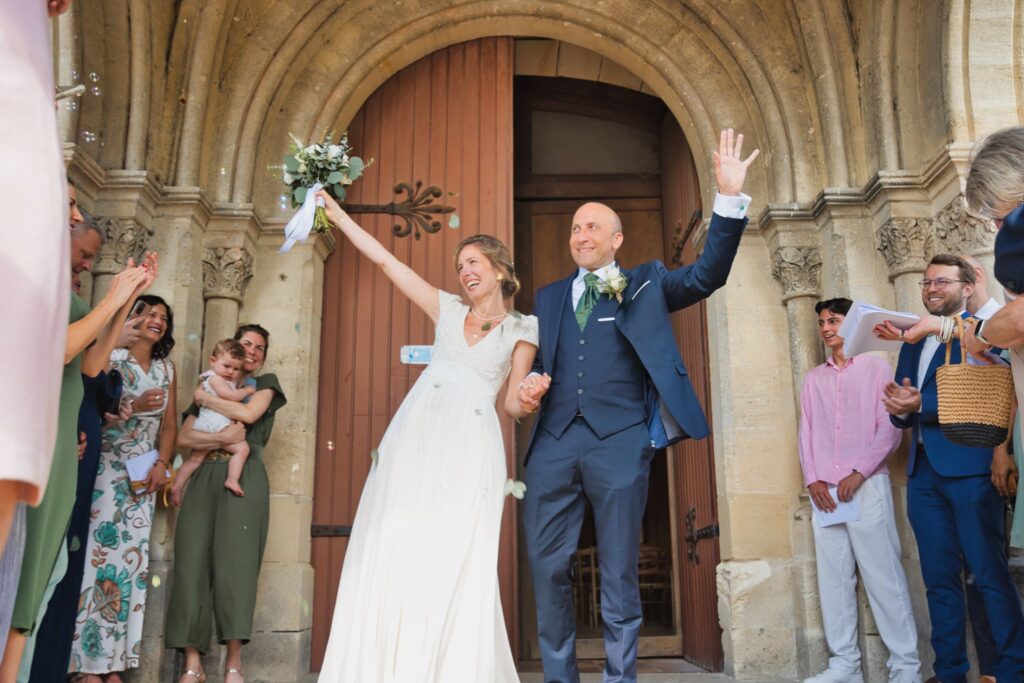 Les mariés sortent de l'église par la porte en bois, un bras levé, entourés d'invités.