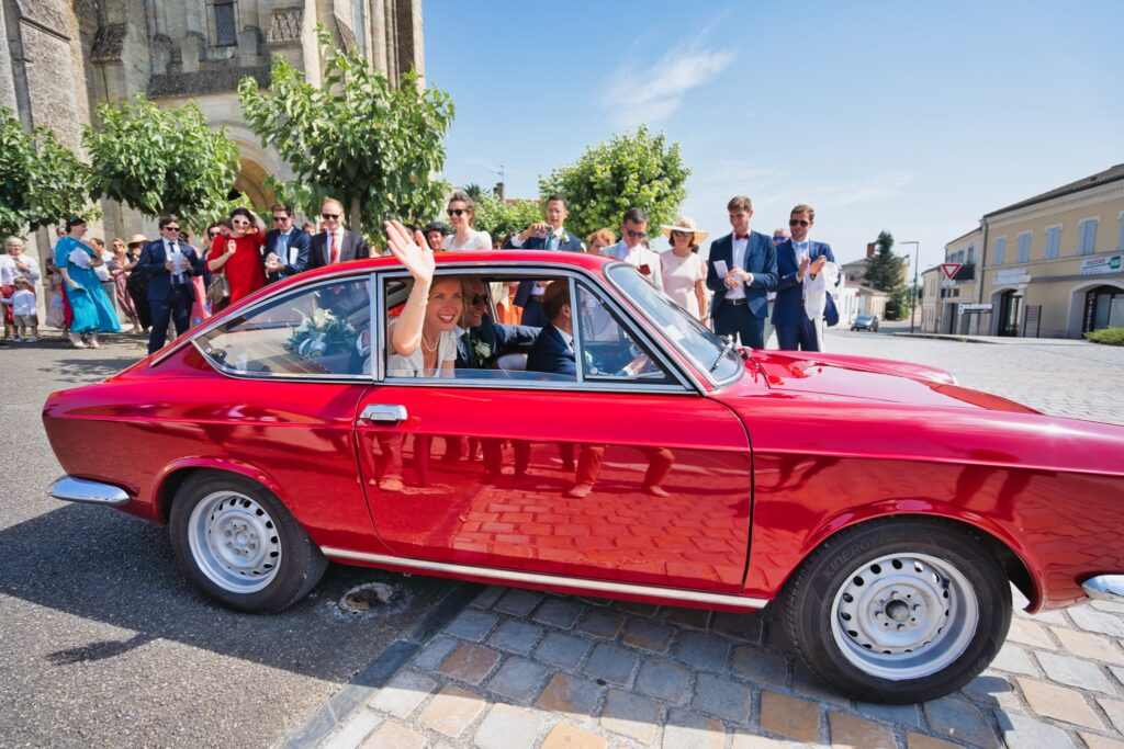 Une voiture ancienne rouge sur une place pavée, avec les mariés à bord et des invités autour.