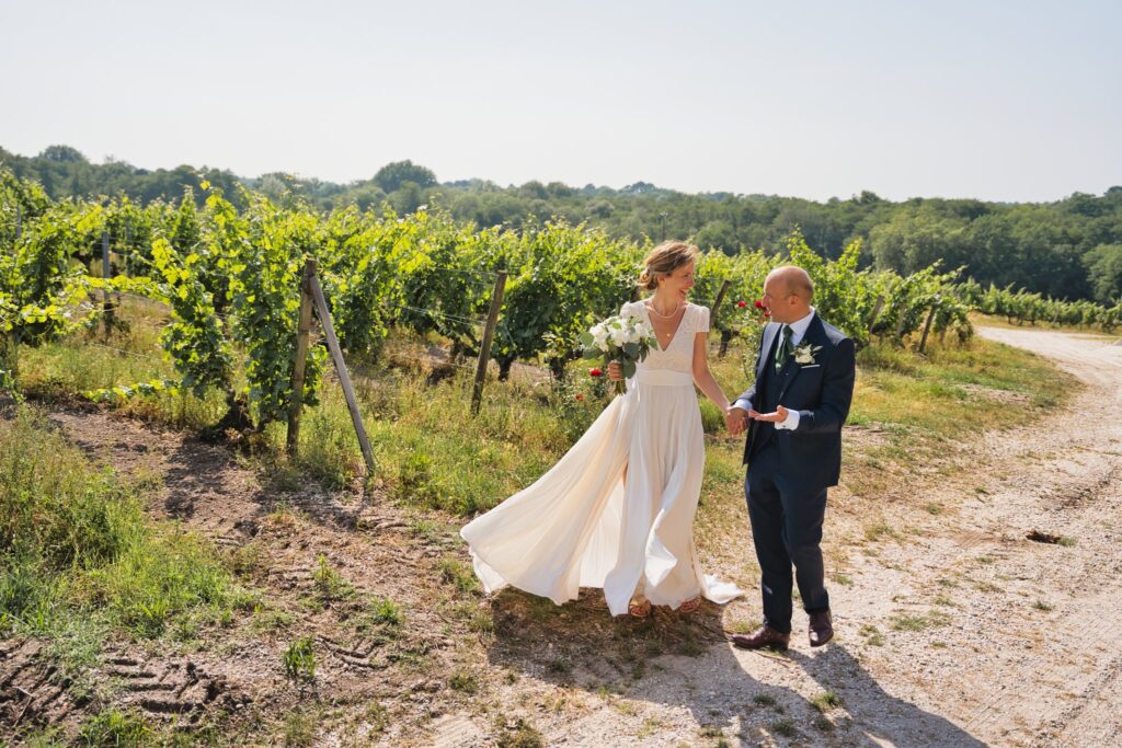 Les mariés sur un chemin de terre entre les rangs de vigne, la robe soulevée par le vent.
