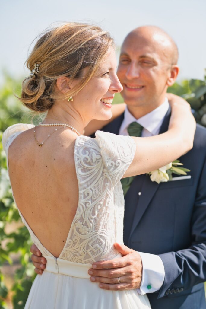 Gros plan sur les mariés enlacés dans les vignes, la mariée de dos riant, la robe ornée de dentelle.