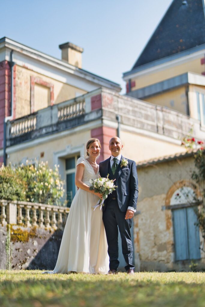 Les mariés debout sur l'herbe devant une façade en pierre, le bouquet dans les mains de la mariée.