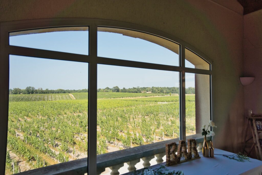 Une grande fenêtre cintrée qui ouvre sur les vignes vertes, avec une enseigne bar en bois au premier plan.