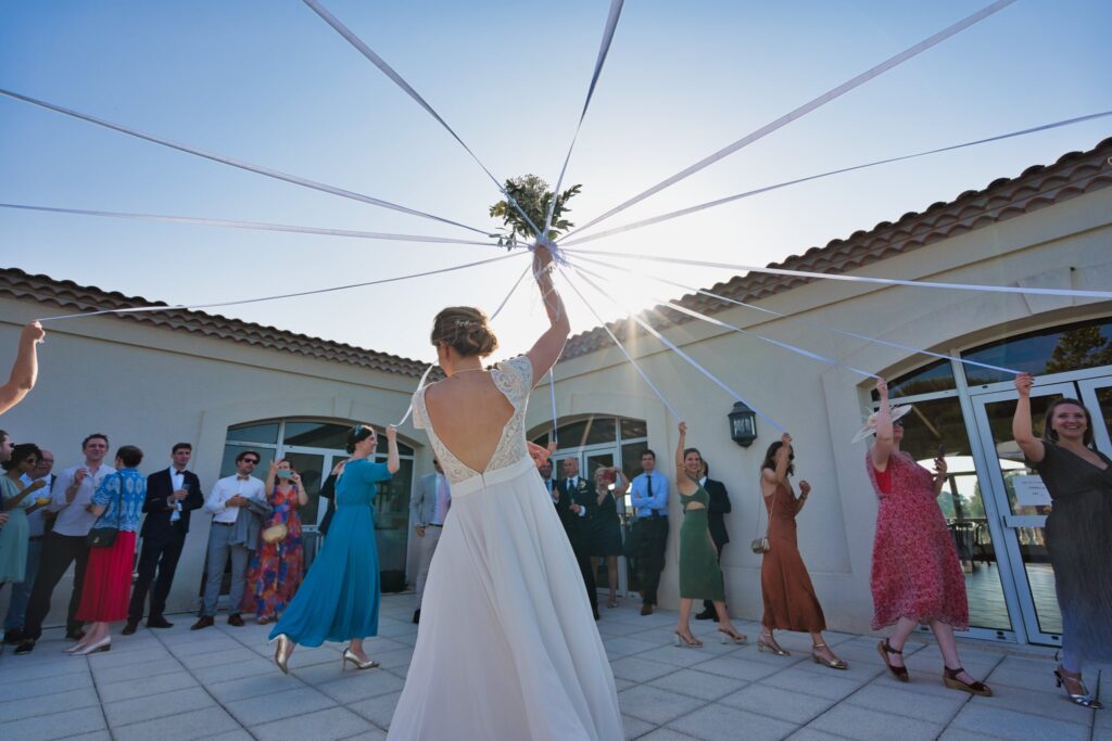 La mariée de dos, bras levé avec son bouquet, au moment du lancer, face aux invités sur la terrasse.