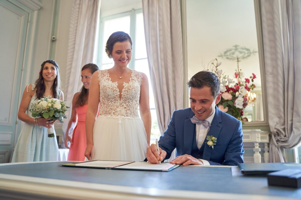 Le marié signe le registre à une table, la mariée debout à côté de lui devant de grandes fenêtres.