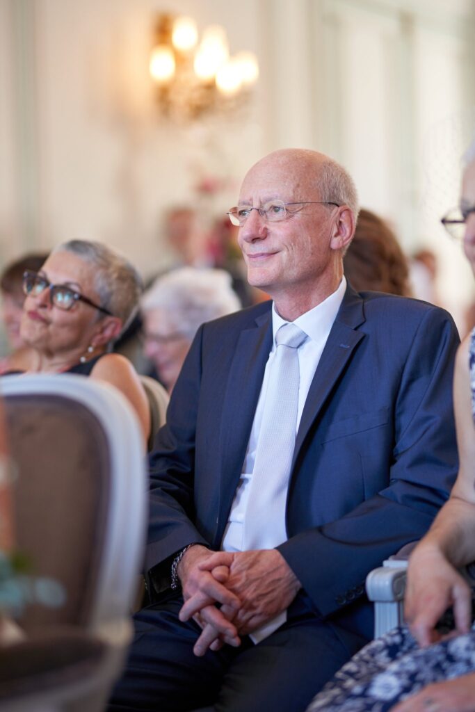 Un homme aux cheveux gris et à lunettes, en costume bleu, assis parmi les invités, les mains jointes.