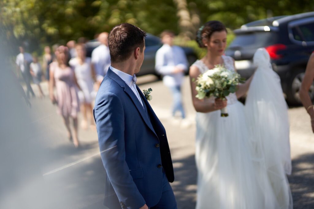 Le marié et la mariée, bouquet à la main, dehors, avec les invités qui marchent derrière eux.