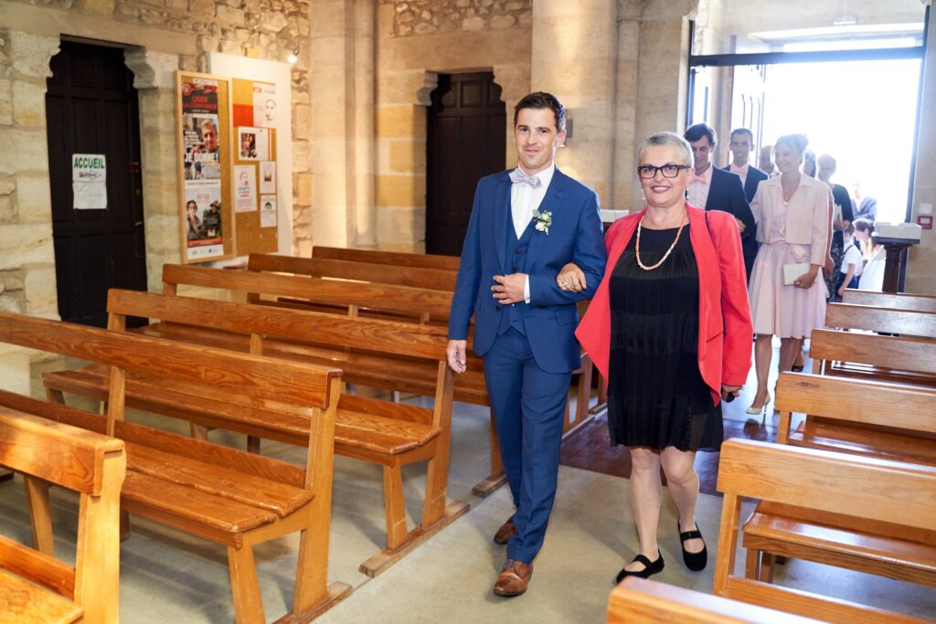 Le marié entre dans l'église au bras d'une femme en veste rouge, entre les bancs en bois.