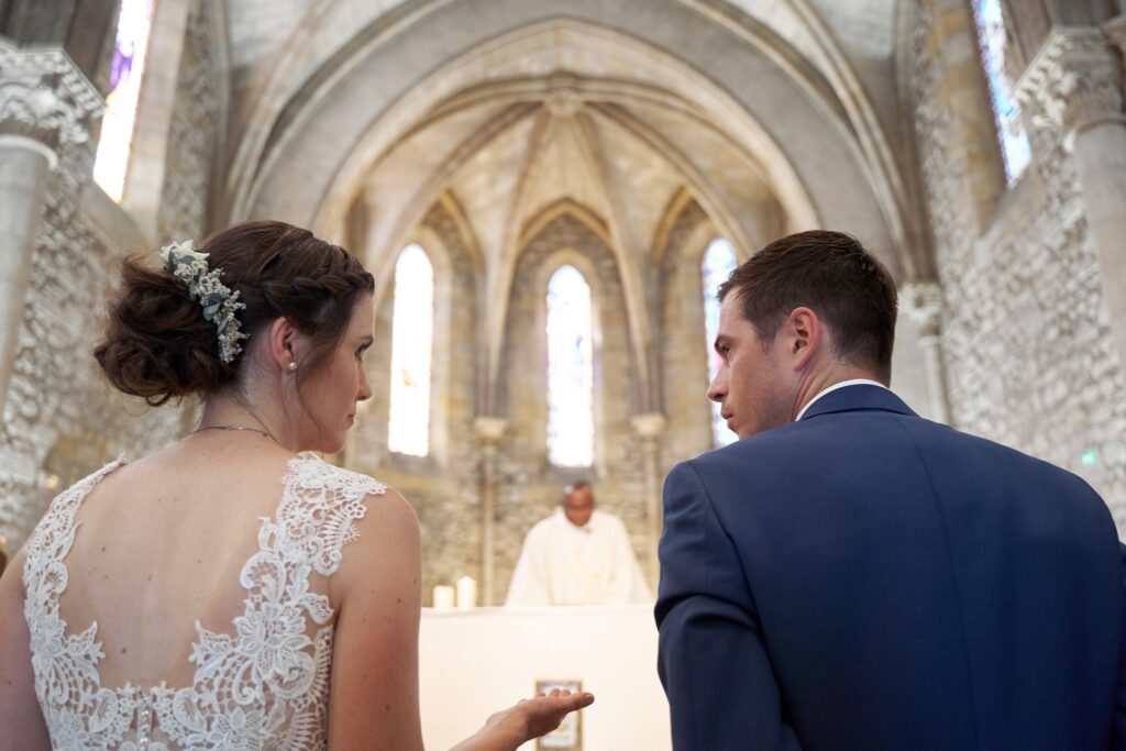 Les mariés côte à côte face au prêtre, sous les fenêtres en ogive de l'église en pierre.