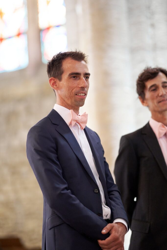 Deux hommes en costume sombre et nœud papillon rose, souriants, dans l'église.