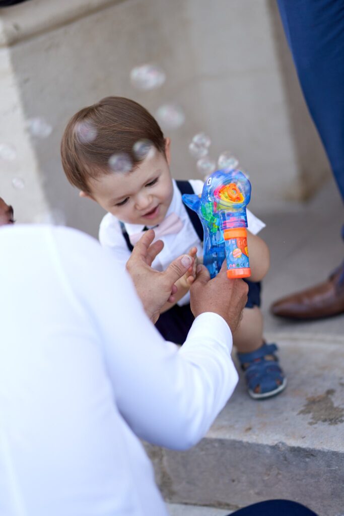 Un petit garçon joue avec un pistolet à bulles de savon, tenu par un adulte en chemise blanche.