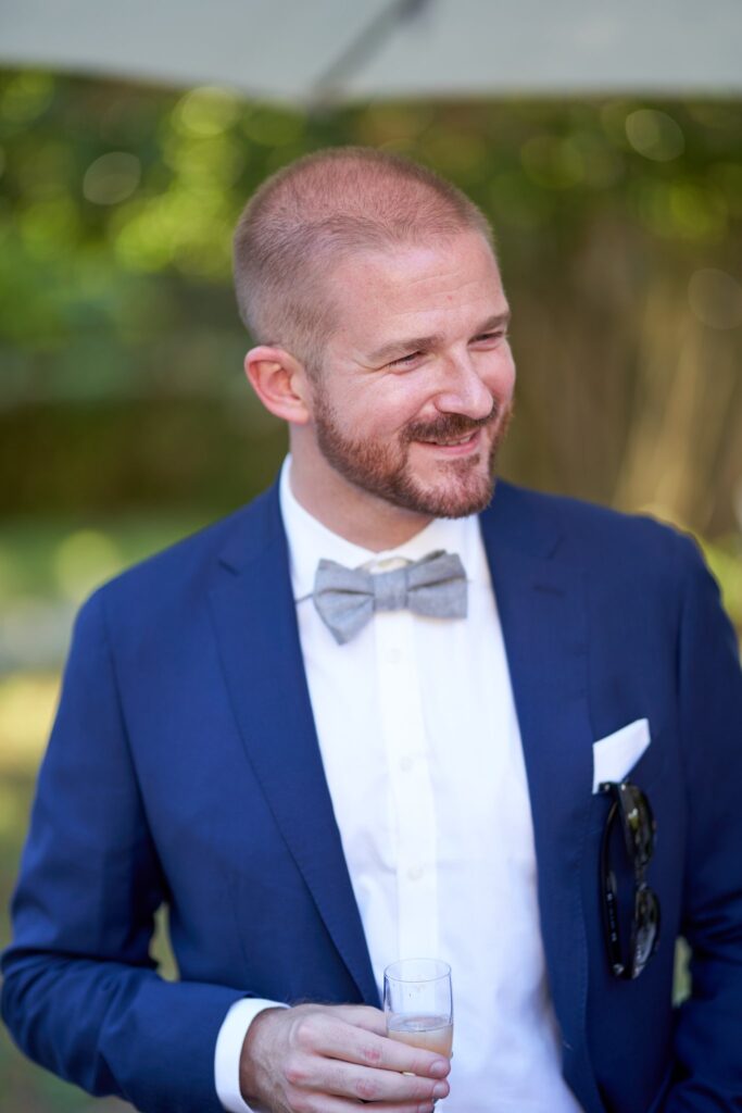 Un homme souriant en costume bleu et nœud papillon gris, un verre à la main, dans un jardin.