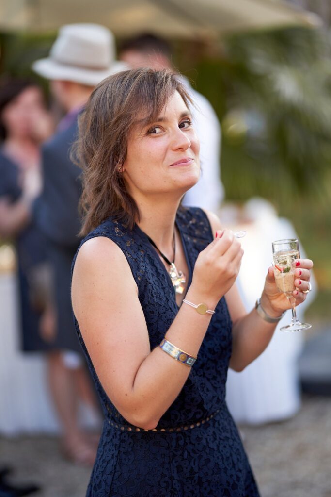 Une femme en robe bleu marine, un verre à la main, devant un groupe d'invités.