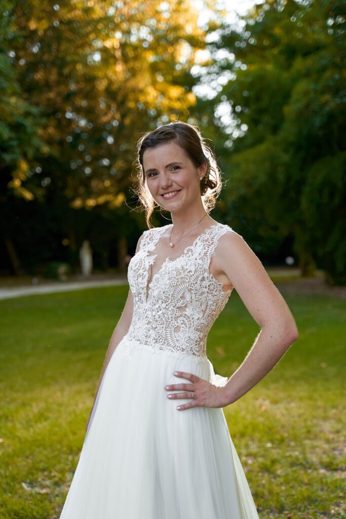 La mariée, une main sur la hanche, debout sur une pelouse devant des arbres éclairés par le soleil bas.