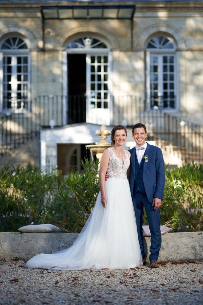 Les mariés sourient côte à côte devant la façade d'un château et une fontaine, dans la lumière dorée de fin de journée.
