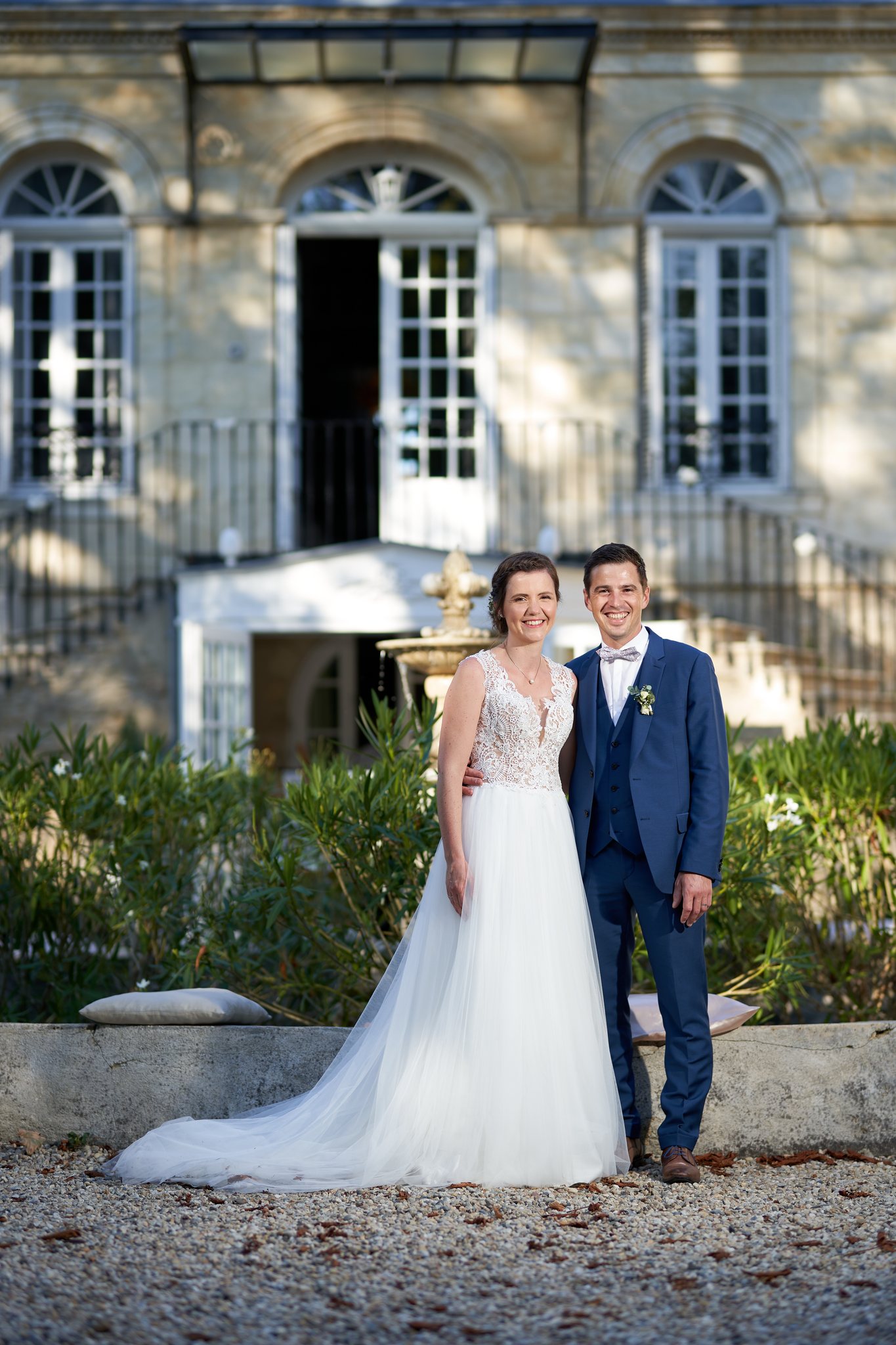 Les mariés sourient côte à côte devant la façade d'un château et une fontaine, dans la lumière dorée de fin de journée.
