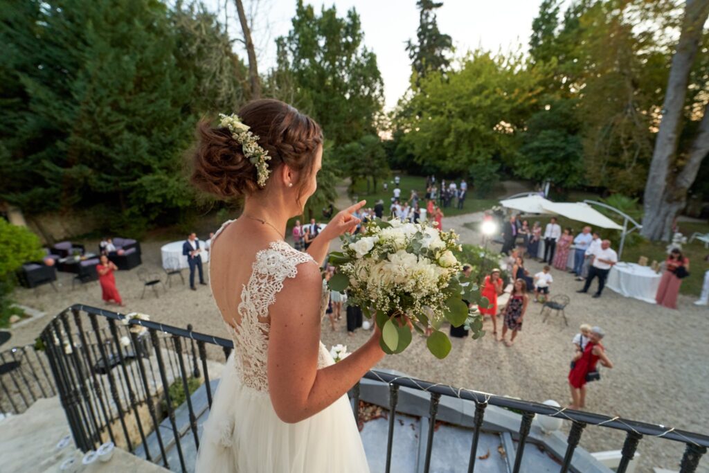 La mariée, bouquet en main, regarde depuis un escalier le jardin où des invités se retrouvent.