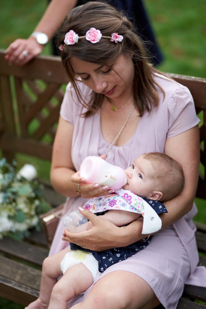 Une jeune femme couronnée de fleurs tient un bébé qui prend le biberon, assise sur un banc en bois.