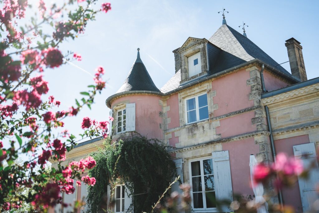 Façade rose du château de Loudenne avec sa tourelle, encadrée par des fleurs rouges, sous un ciel bleu