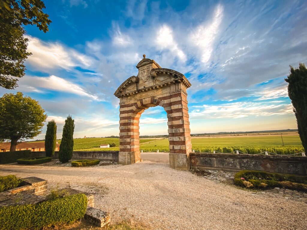 Portail monumental en pierre et brique ouvert sur les vignes sous un ciel dégagé, en fin de journée