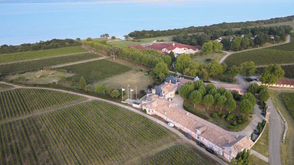 Vue aérienne du château et de ses dépendances au milieu des vignes, avec l'estuaire de la Gironde à l'horizon