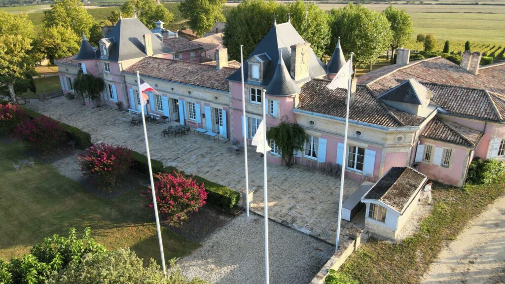 Vue aérienne de la façade rose du Château Loudenne au soleil, avec ses drapeaux, ses massifs de fleurs rouges et la terrasse