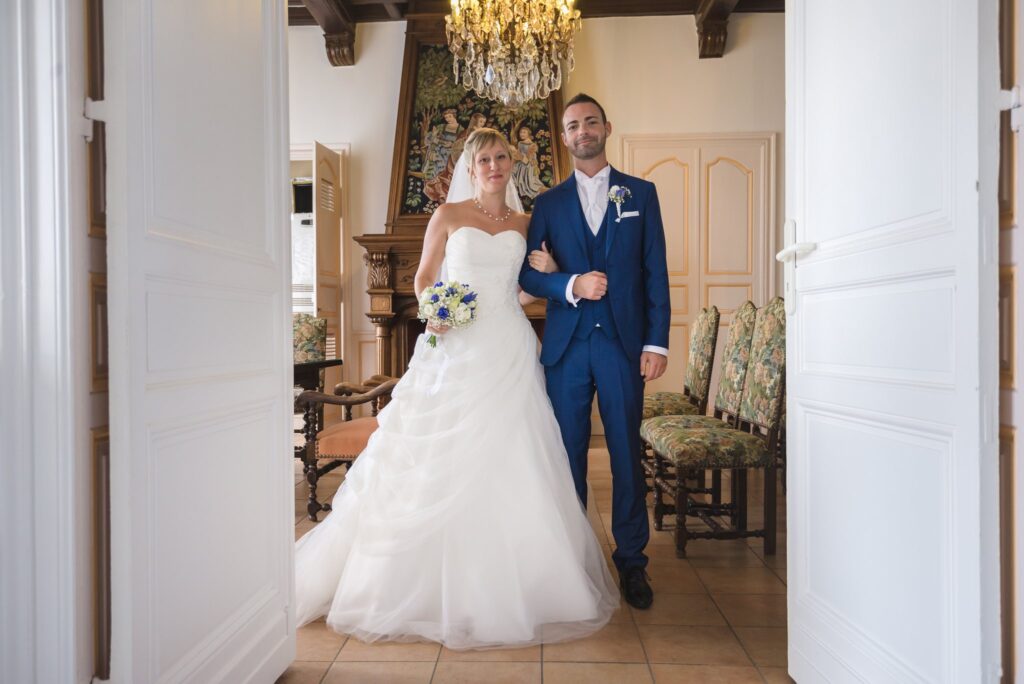 La mariée et le marié bras dessus bras dessous dans l'encadrement de grandes portes blanches, sous un lustre.