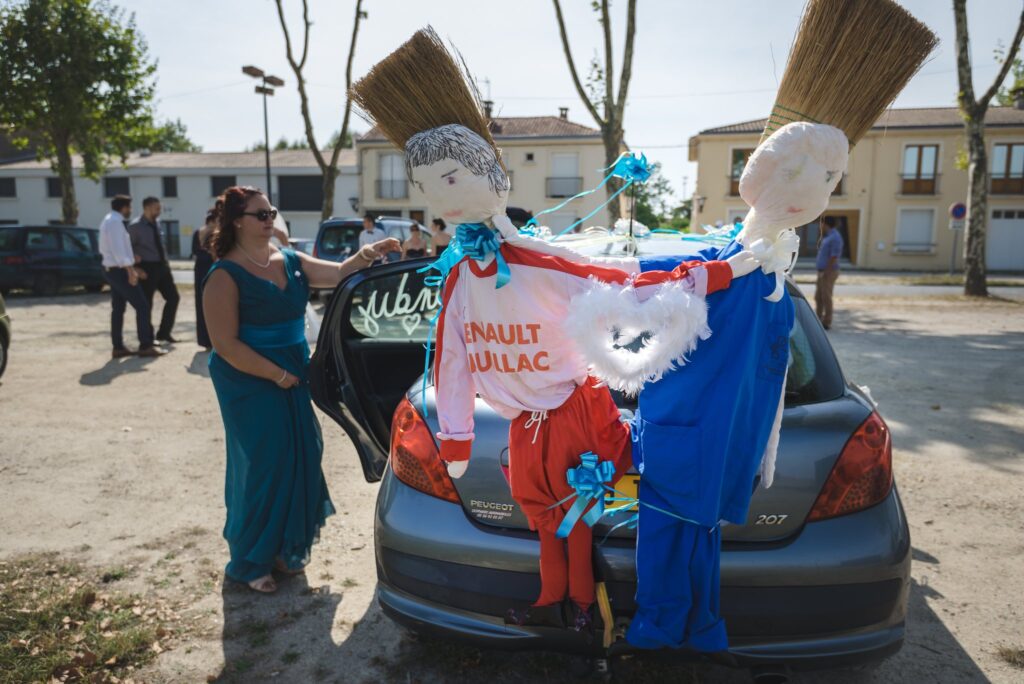 Une voiture décorée à l'arrière de deux poupées en tissu aux couleurs bleu, blanc et rouge, sur une place ensoleillée.