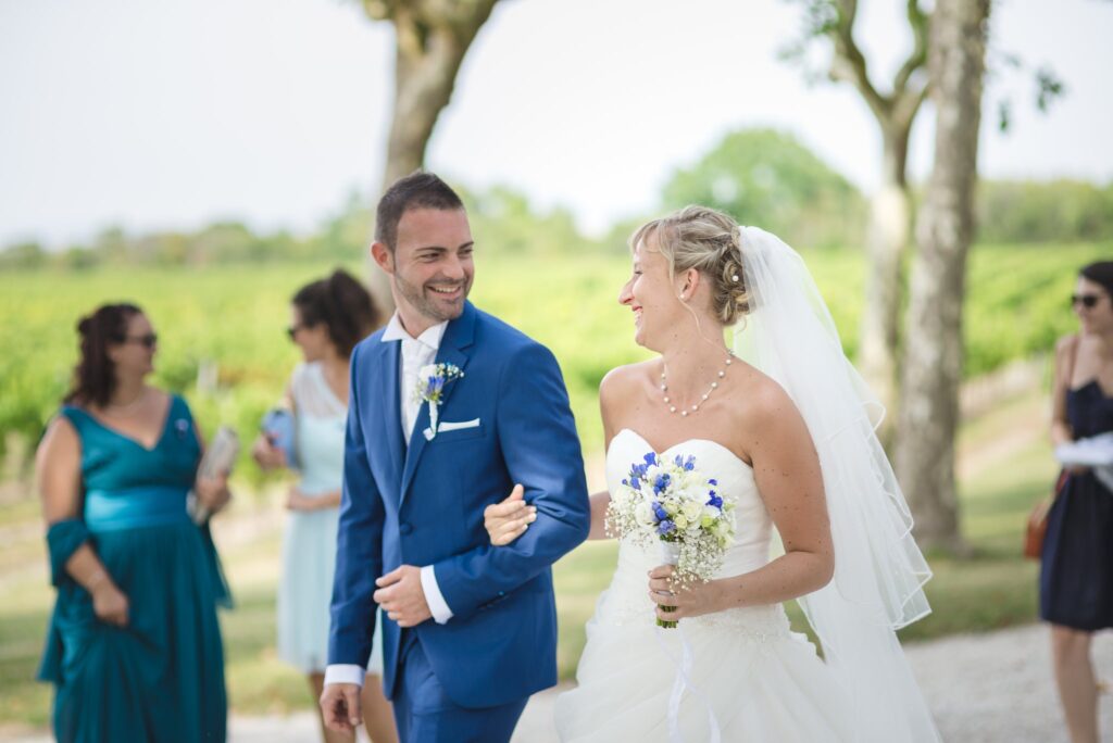 Le marié et la mariée se sourient en marchant au bras l'un de l'autre, avec des vignes et des invités derrière eux.