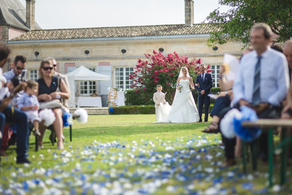 La mariée avance dans l'allée de pétales bleus et blancs, entre deux rangées d'invités assis, devant la façade du château.