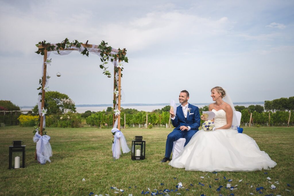 Les mariés assis sous une arche de bois ornée de feuillage, sur une pelouse bordée de vignes, avec deux lanternes au sol.