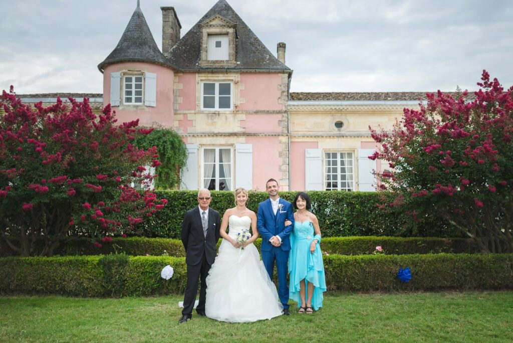 Les mariés posent entre un homme et une femme devant la façade rose du château, encadrée de massifs fleuris rouge vif.