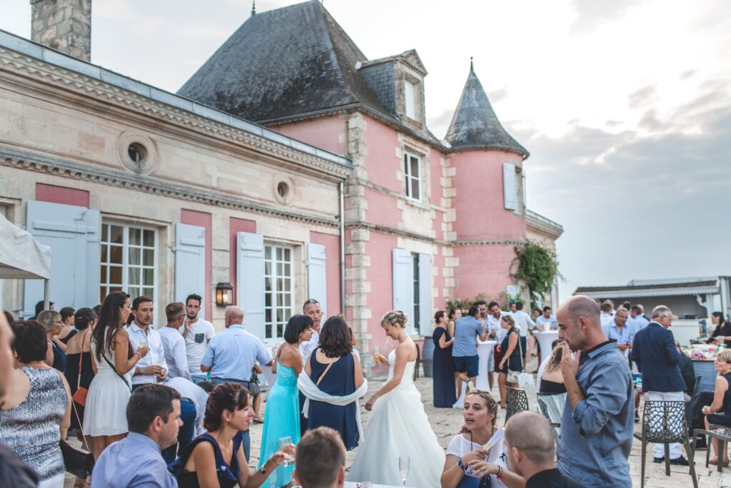 Des invités debout sur la terrasse du château, verre à la main, avec la façade rose et ses tourelles en arrière-plan.