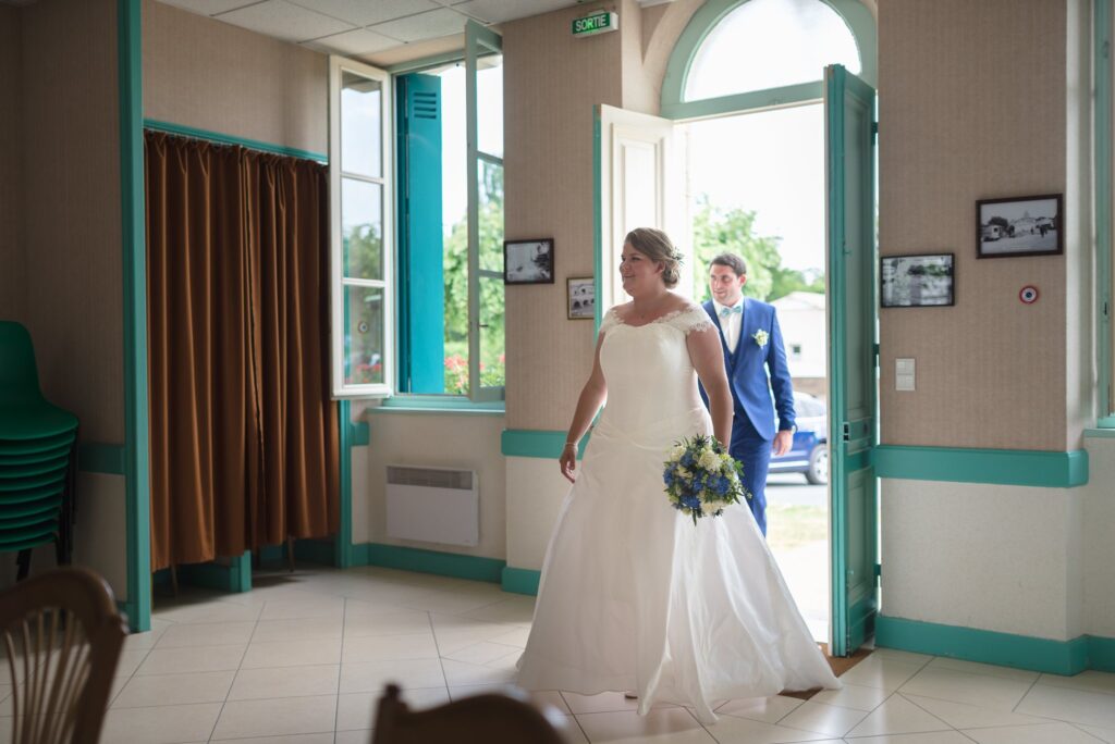 Une mariée en robe blanche avançant dans la salle de la mairie, un bouquet à la main.
