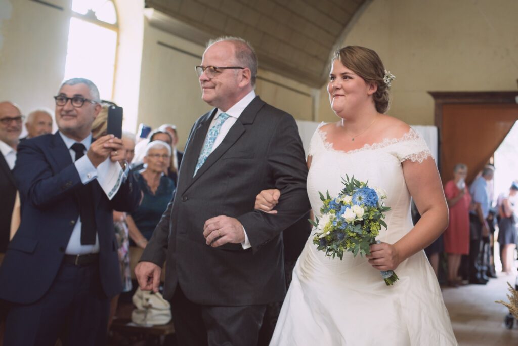 Un homme donne le bras à la mariée qui avance dans l'allée de l'église, entourés d'invités.