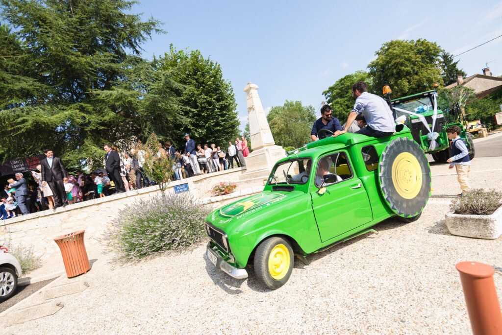 Une 4L peinte en vert avec une roue de tracteur, devant un tracteur décoré de rubans et une foule d'invités.