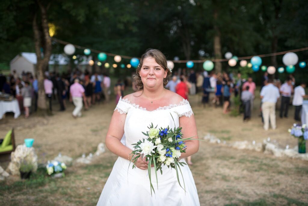 La mariée sourit en tenant son bouquet blanc et bleu dans le parc, devant une guirlande de lampions colorés et des invités flous.