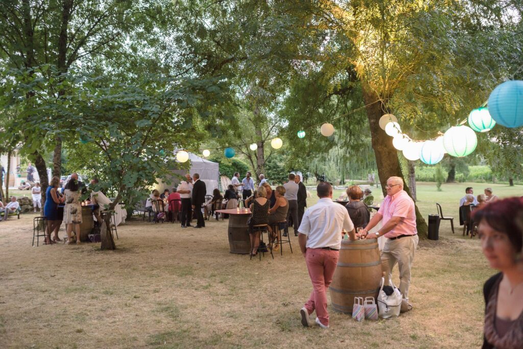 Des invités réunis sous les arbres autour de tonneaux, éclairés par des guirlandes et des lampions.