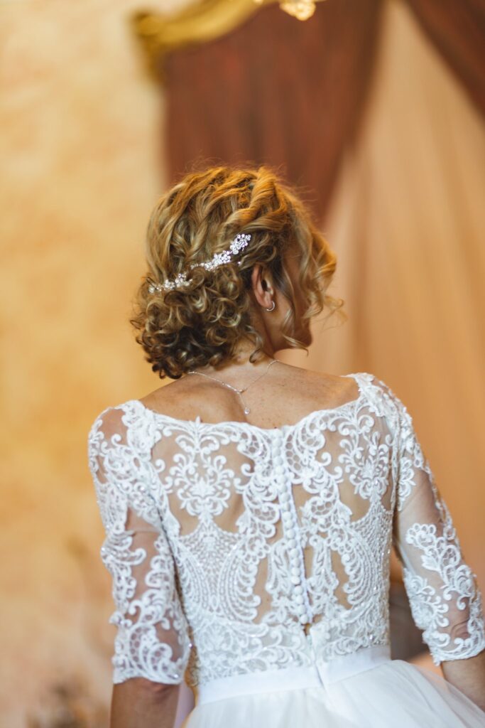 Une mariée vue de dos, le haut de sa robe en dentelle et sa coiffure ornée de petits bijoux.