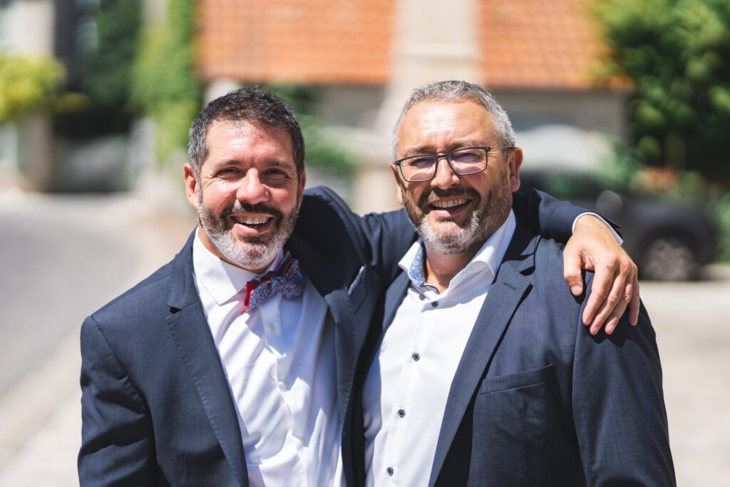 Deux hommes en costume sourient en s'enlaçant les épaules dans une rue.