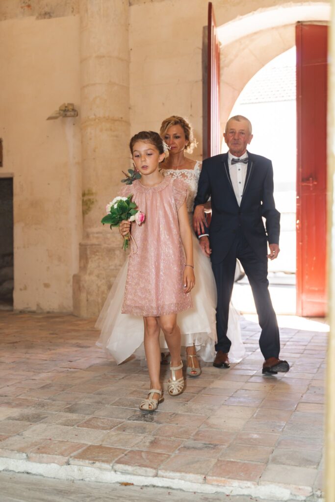 Un homme en costume et une mariée entrent dans l'église, accompagnés d'une petite fille tenant un bouquet.
