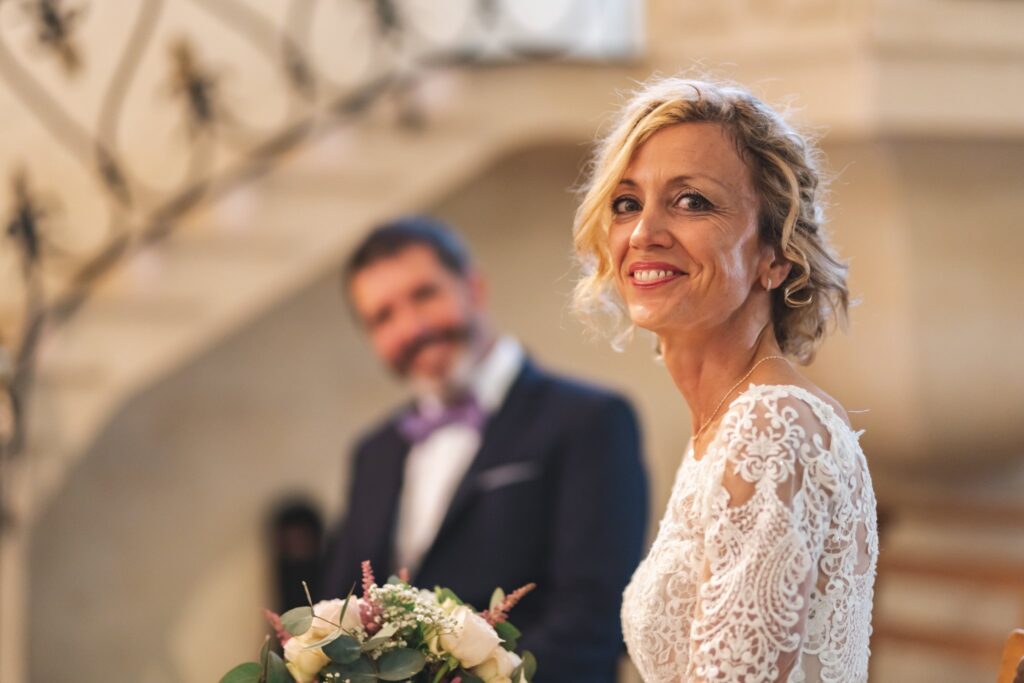 Une mariée souriante en robe de dentelle tient son bouquet, le marié flou derrière elle devant un escalier en fer forgé.