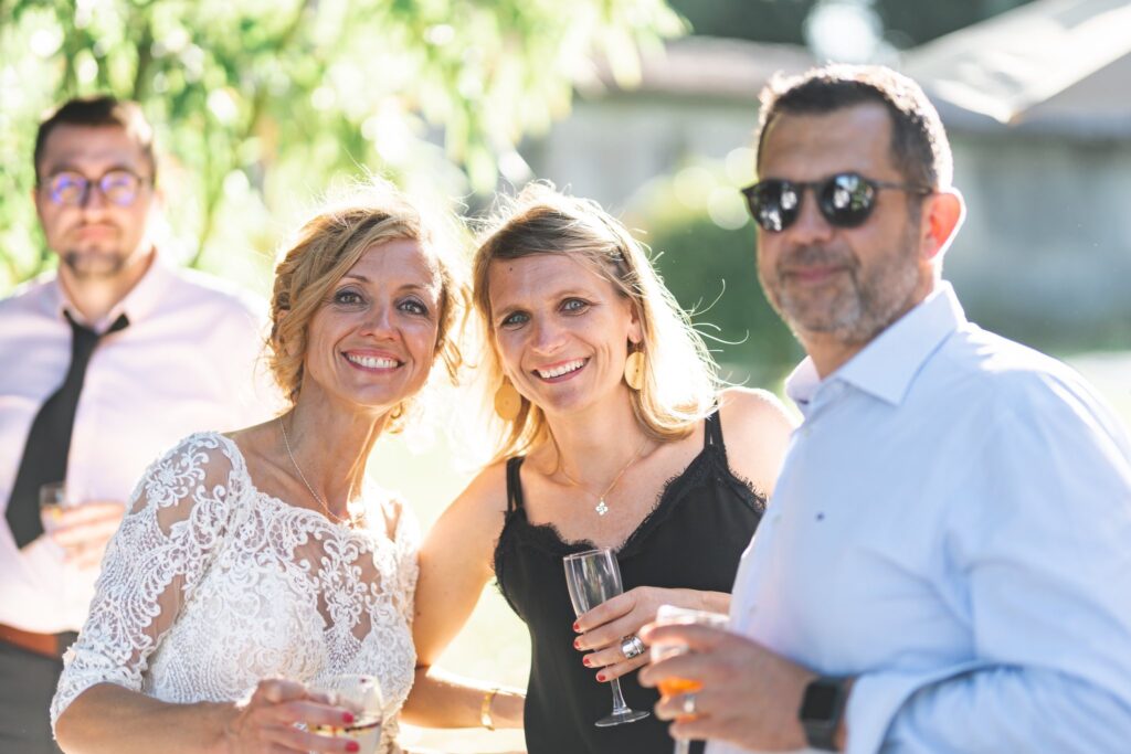 Une mariée et des invités souriants, verre à la main, en extérieur.