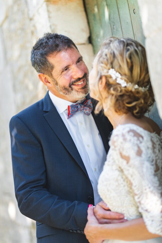Le marié et la mariée enlacés se sourient, adossés à un mur de pierre.