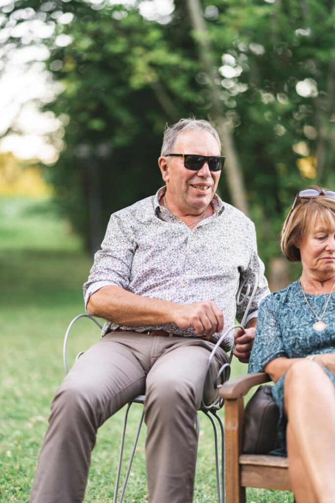 Un homme à lunettes de soleil et une femme assis sur des chaises dans l'herbe, sous les arbres.