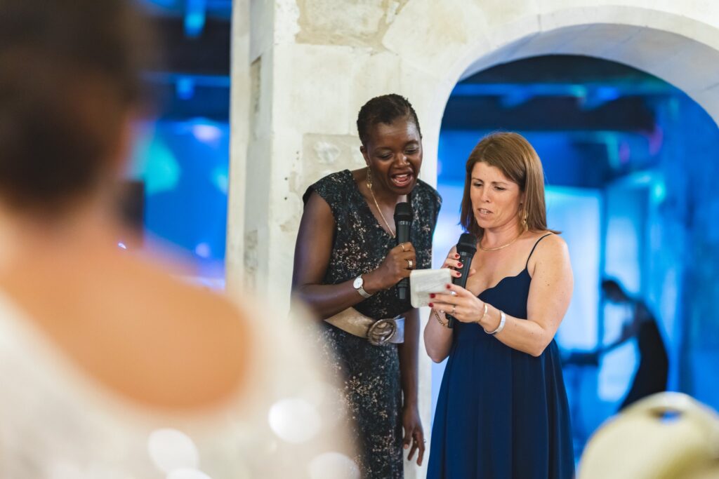 Deux femmes parlent au micro sous une arche éclairée de bleu, la mariée au premier plan, de dos.